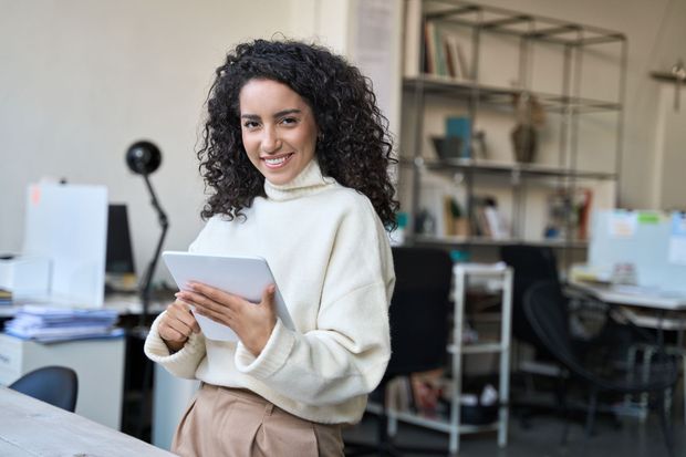 Mujer con cabello rizado, sonriendo, sosteniendo una tableta en el entorno de oficina.