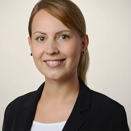 Frau in schwarzem Blazer lächelt, Pferdeschwanz, vor hellem Hintergrund.