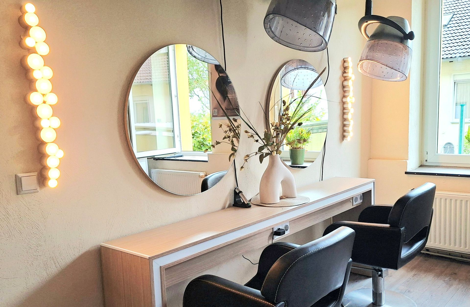 Moderner Salonarbeitsplatz mit zwei Spiegeln, weißer Ablage, schwarzen Stühlen und dekorativer Beleuchtung