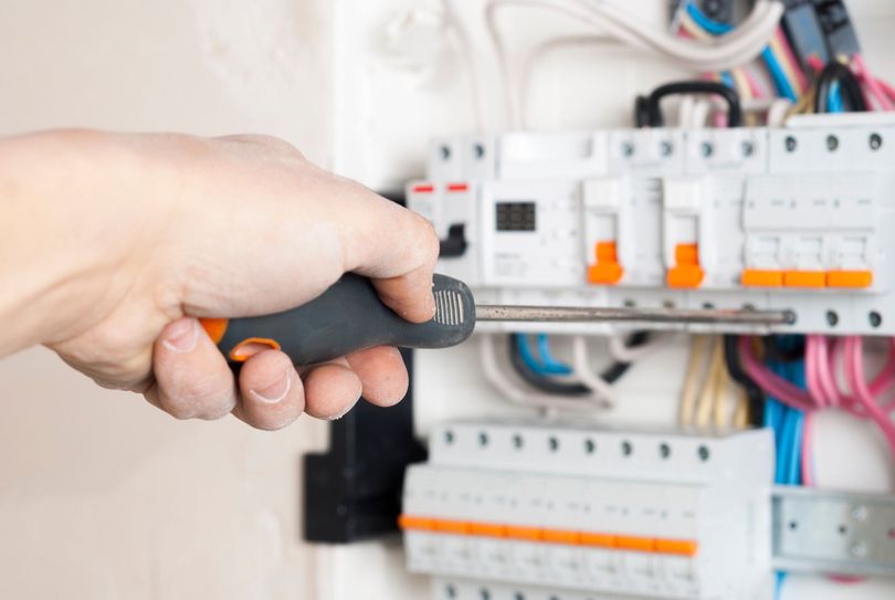 Mano con destornillador trabajando en un panel eléctrico con disyuntores y cableado.