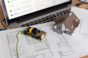 Computadora portátil, modelo de casa, placa de circuito y planos sobre un escritorio, lo que sugiere la integración de tecnología de hogar inteligente.