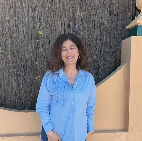 Una mujer de cabello castaño y rizado sonríe con una camisa azul con diseño floral. Está de pie sobre un fondo marrón y amarillo.