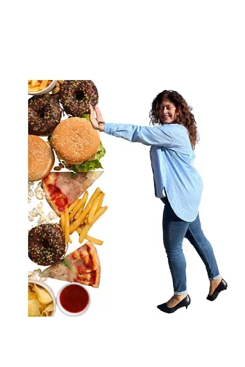 Una mujer aparta una columna de comida chatarra: donas, hamburguesas, papas fritas, pizza y patatas fritas. Lleva una camisa azul, jeans y tacones, sonriendo.