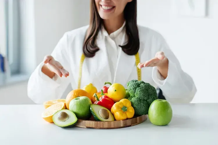 Dietista presentando una bandeja de madera con frutas y verduras de colores, sonriendo en una habitación llena de luz.