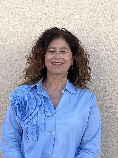 Una mujer de cabello castaño rizado sonríe, vistiendo una camisa azul claro con un gran detalle de flores en el hombro, de pie frente a una pared beige.