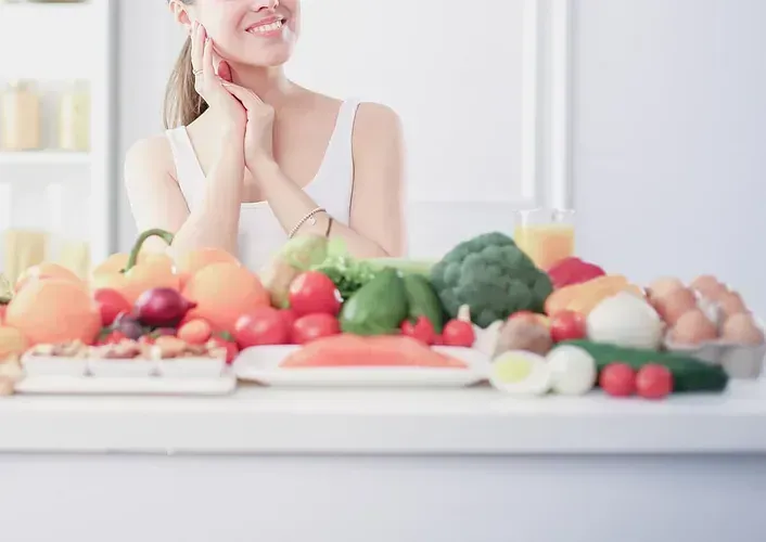 Mujer sonriendo, con los brazos cerca de la cara, de pie detrás de una mesa cargada de frutas frescas, verduras y huevos en una cocina luminosa.
