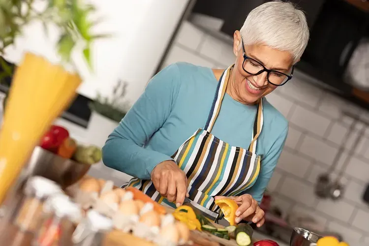 Una mujer con cabello corto gris y gafas sonríe mientras corta verduras en una cocina, vistiendo una camisa azul y un delantal de rayas.
