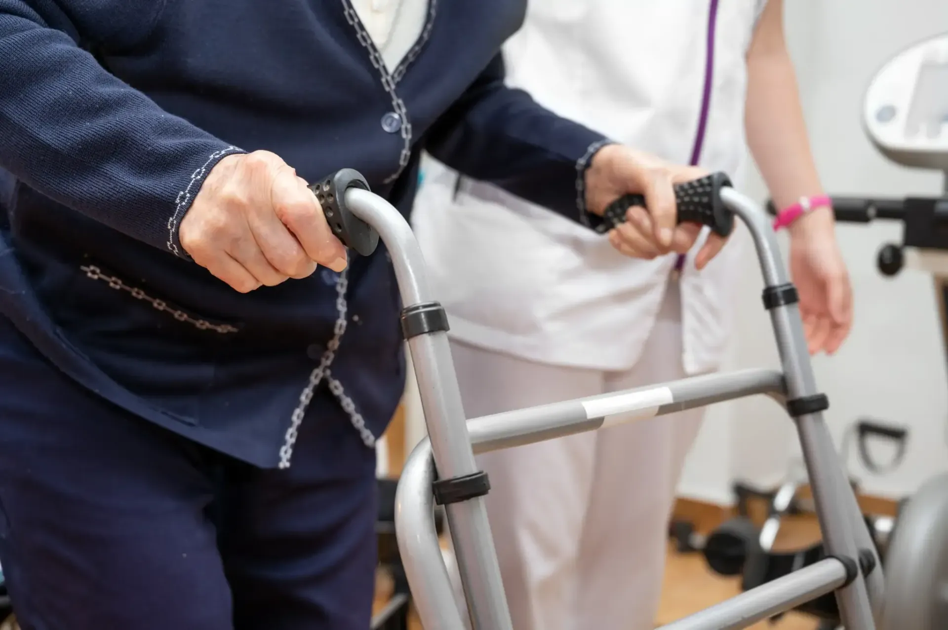 Una persona mayor usa un andador, asistida por una persona con uniforme blanco. En el interior.