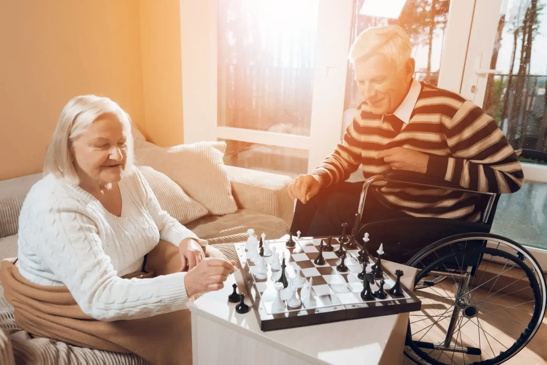 Pareja de ancianos jugando al ajedrez en el interior; hombre en silla de ruedas, sonriendo.