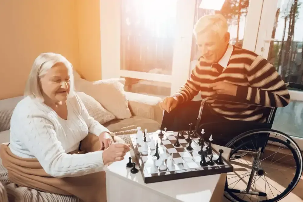 Pareja de ancianos jugando al ajedrez en el interior; hombre en silla de ruedas, sonriendo.