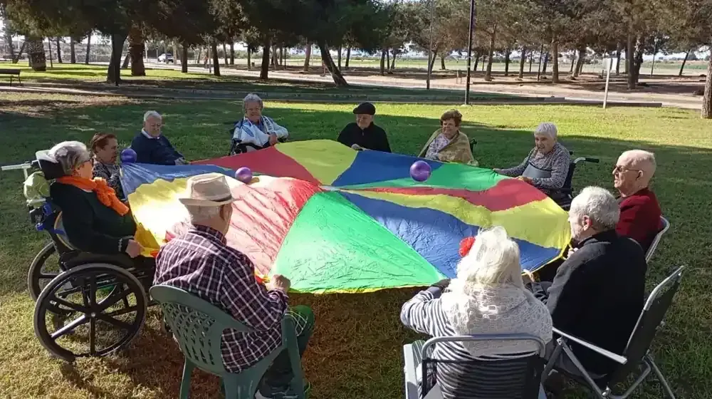 Grupo de personas sentadas alrededor de un paracaídas colorido al aire libre, sujetando los bordes.