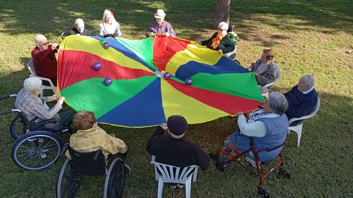 Un cuidador con uniforme rosa revisa documentos con un hombre en silla de ruedas; están en una sala de estar.