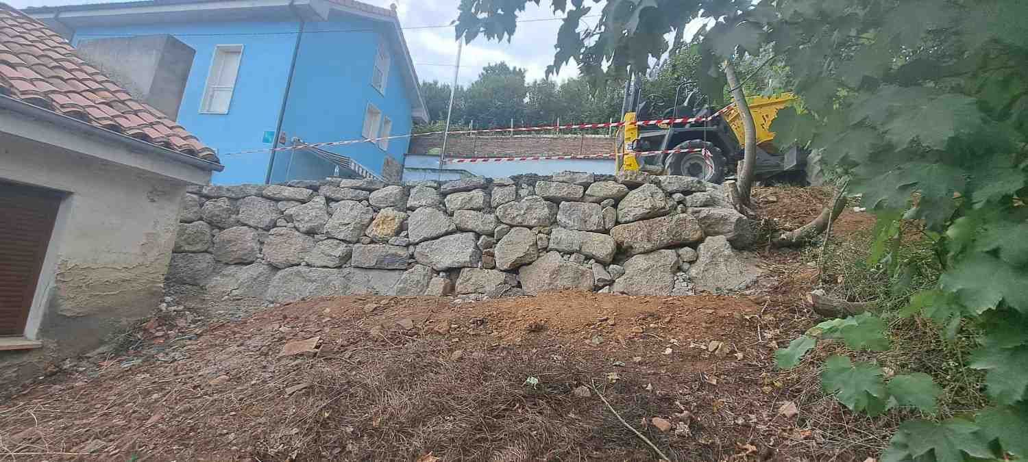 Vehículo de construcción amarillo sobre un muro de contención de piedra junto a una casa azul y una pendiente de tierra.