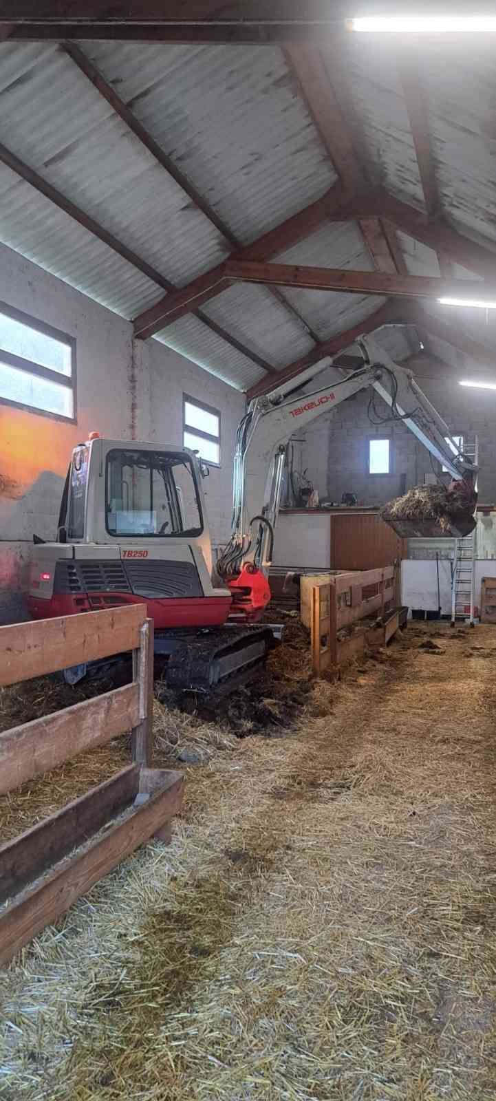 Interior de un cobertizo agrícola con un tractor rojo, otros equipos y suelo cubierto de paja bajo un techo metálico alto.