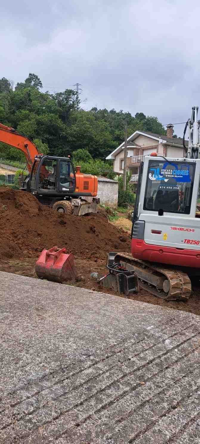 Excavadora y pequeña topadora retirando tierra en una pendiente al costado de una carretera junto a una casa y árboles.