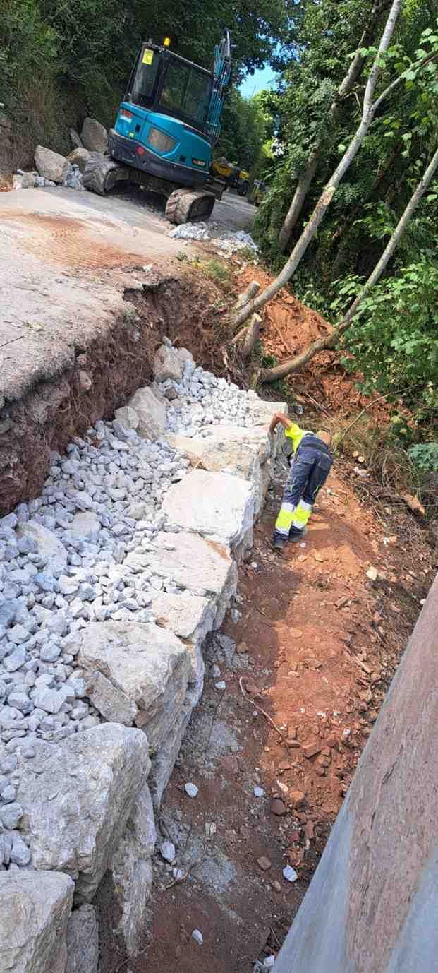 Obras de construcción al borde de la carretera con excavadora, trabajadores y una zanja derrumbada.