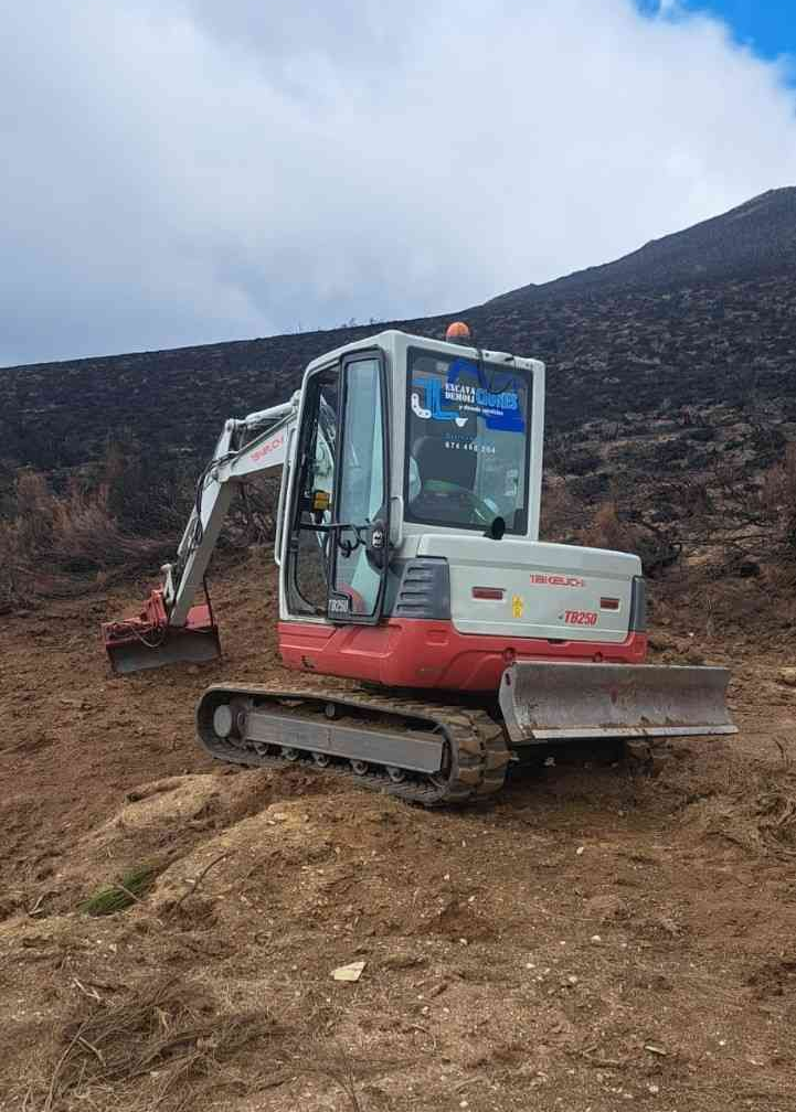 Una pequeña excavadora en una ladera de tierra cerca de montañas rocosas bajo un cielo nublado.