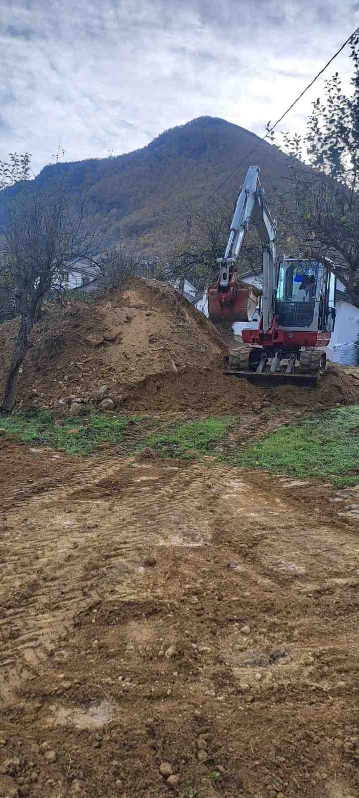 Excavadora despejando un camino de tierra fangosa con una montaña y árboles al fondo.