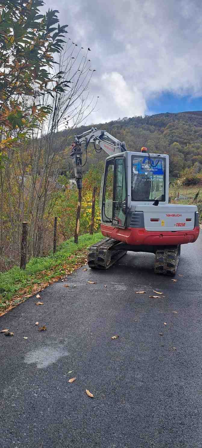 Excavadora roja en un camino rural mojado junto a árboles otoñales y colinas bajo un cielo nublado.