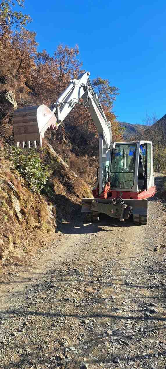 Excavadora en un camino de grava junto a una ladera empinada, excavando en la tierra bajo un cielo azul despejado.