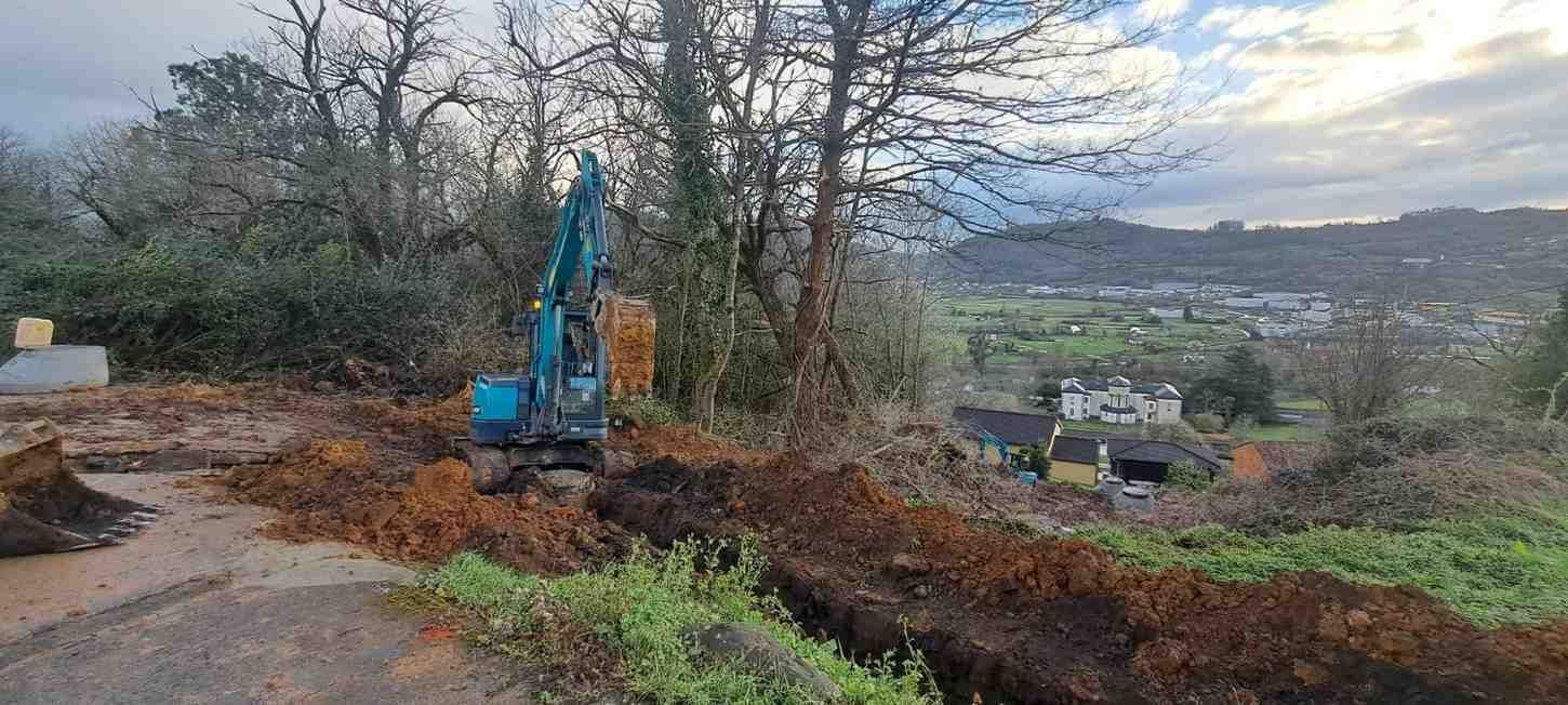 Una excavadora azul excava junto a una ladera fangosa con árboles y una vista del valle al fondo.