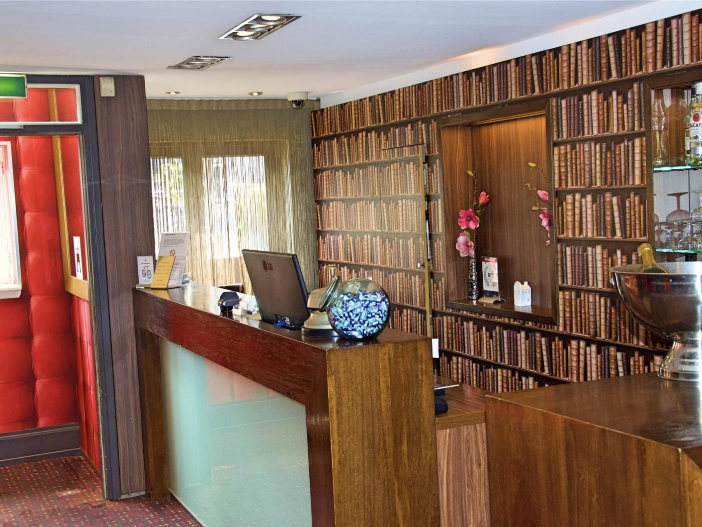 Hotelreceptie met boekenplankbehang. Houten balie en rode, met kussens beklede wand links.