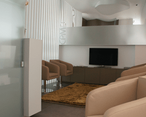 Sala de espera moderna com poltronas bege sobre um tapete dourado, uma televisão sobre um móvel marrom