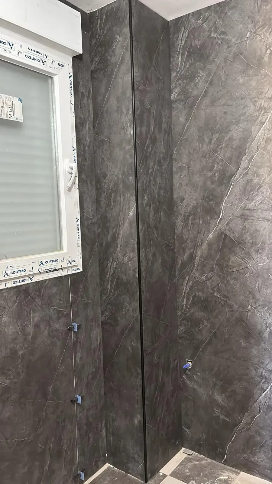Baño con azulejos grises en la esquina, con una grieta vertical alargada. Hay una ventana a la izquierda.