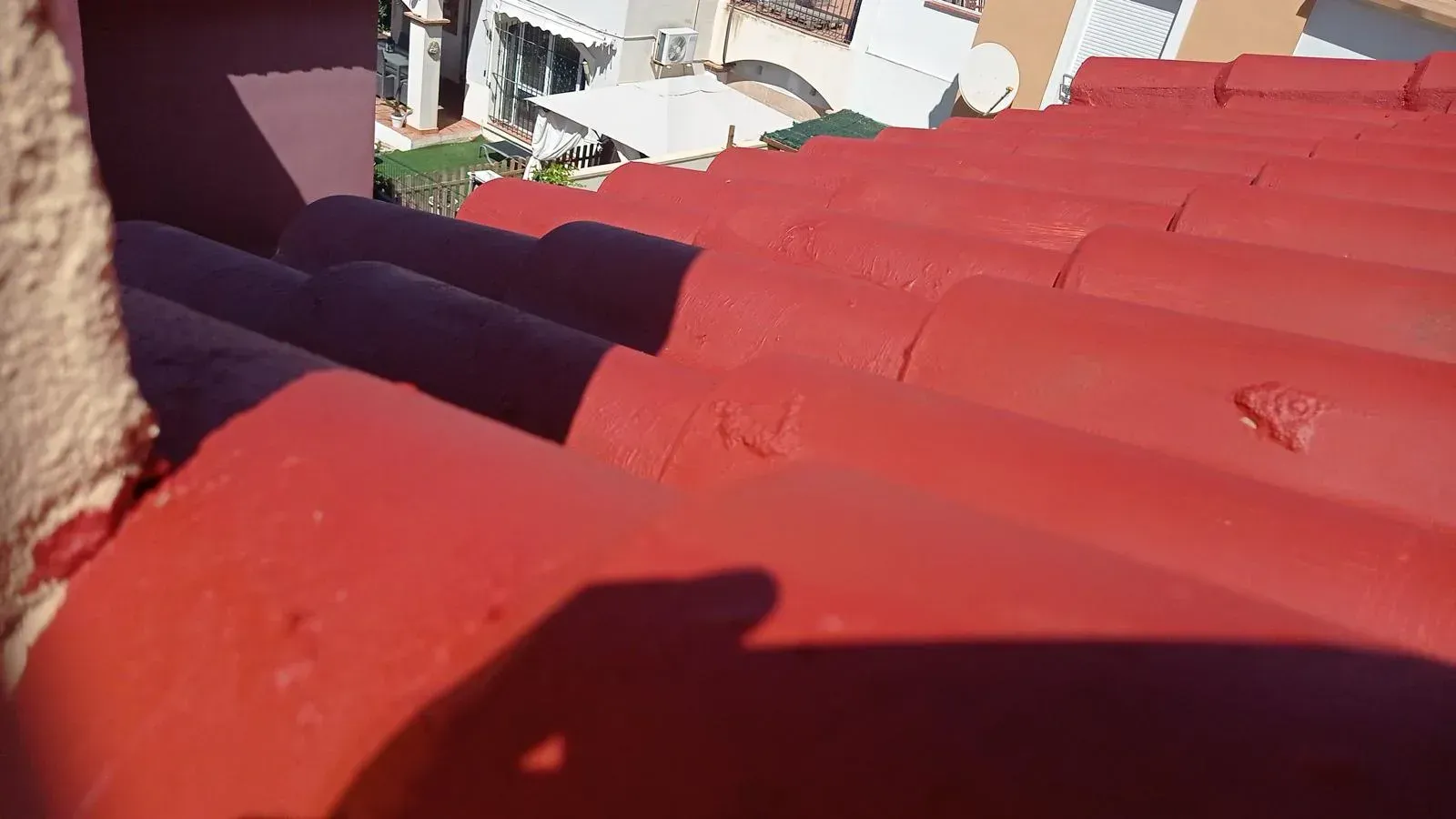 Techo de tejas de barro pintadas de rojo con sombras, con vista a un patio blanco y edificios.