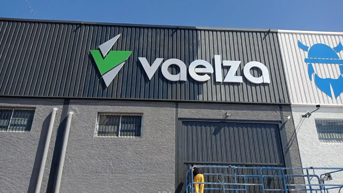 Cartel de Vaelza en un edificio; presenta un logotipo verde y blanco, una fachada gris y negra y un símbolo vikingo azul.