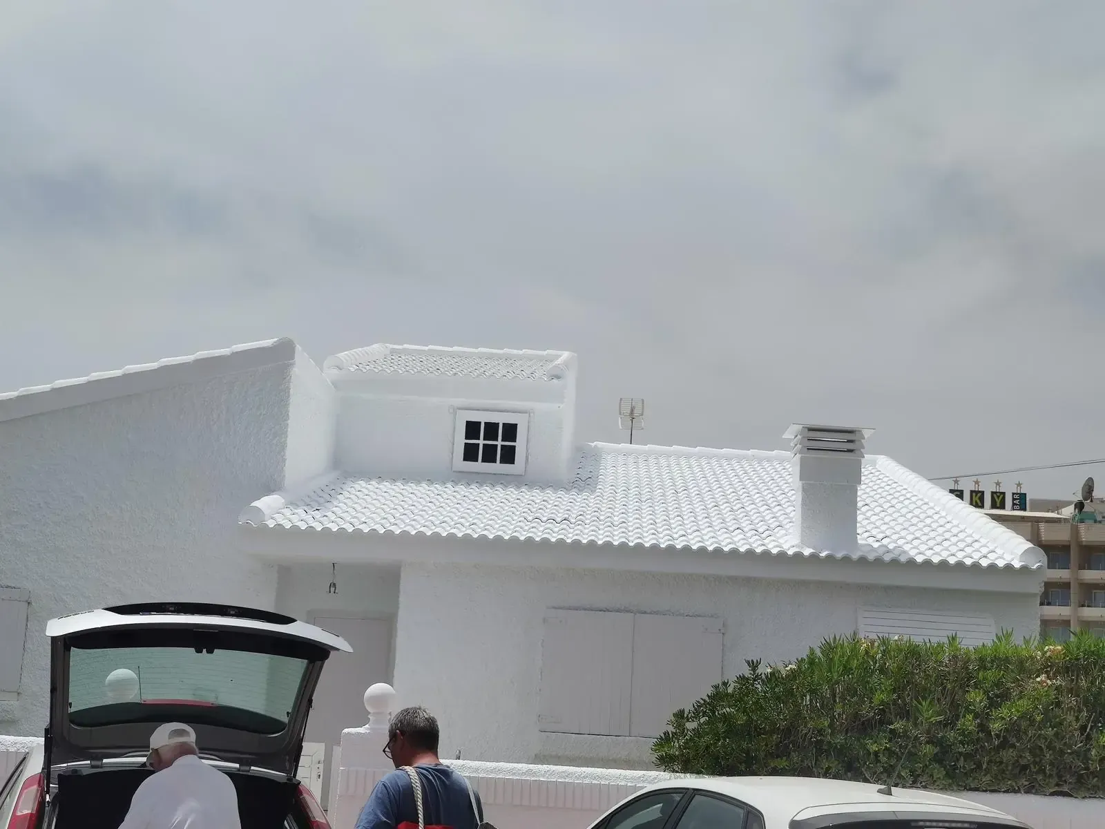 Casa de estuco blanco con maletero de coche abierto y gente, bajo un cielo nublado.