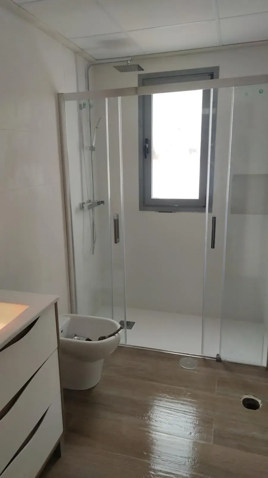 Baño con ducha de cristal, inodoro, lavabo y ventana. Suelo de madera.