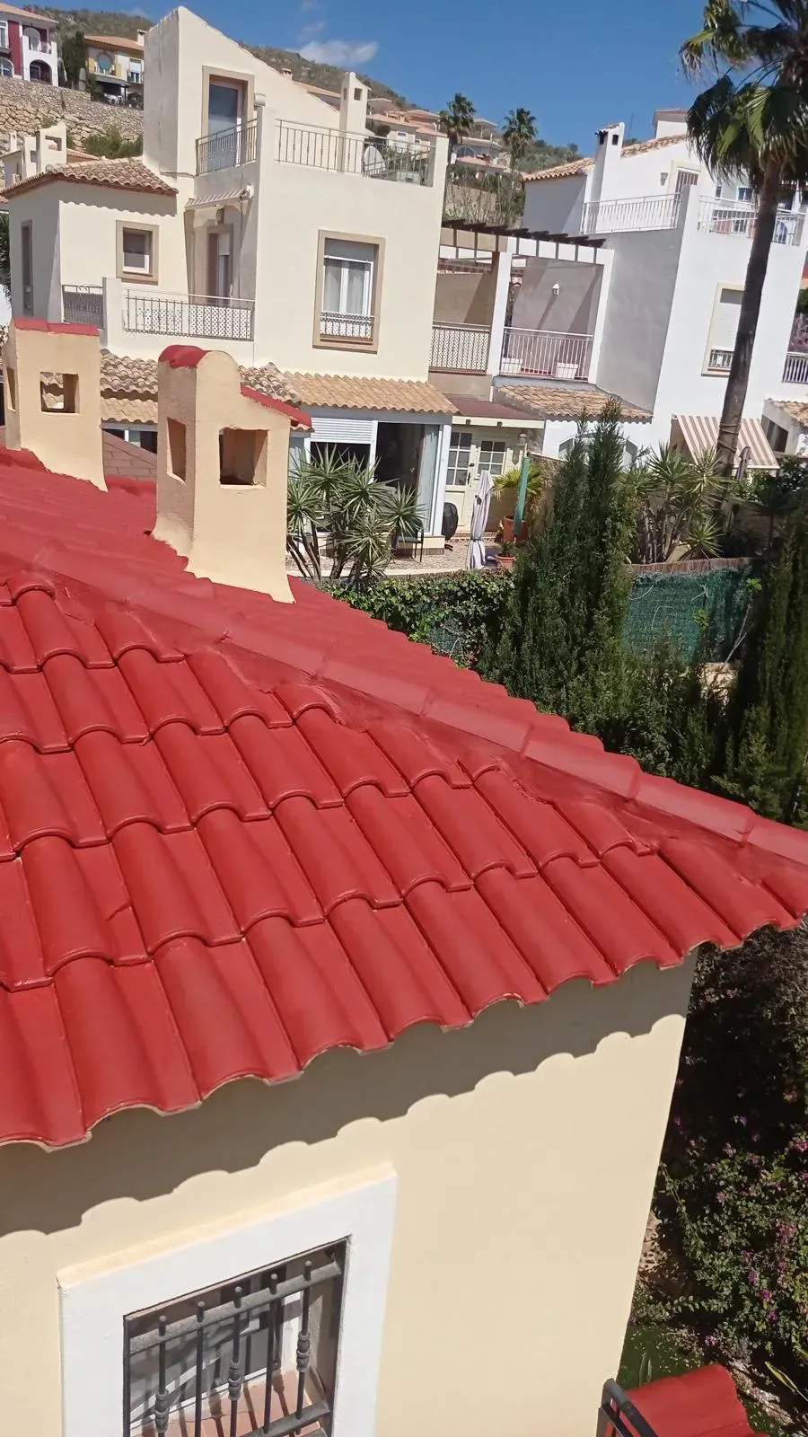 Techo de tejas rojas con dos chimeneas, con vistas a edificios de color crema y árboles verdes bajo un cielo azul.
