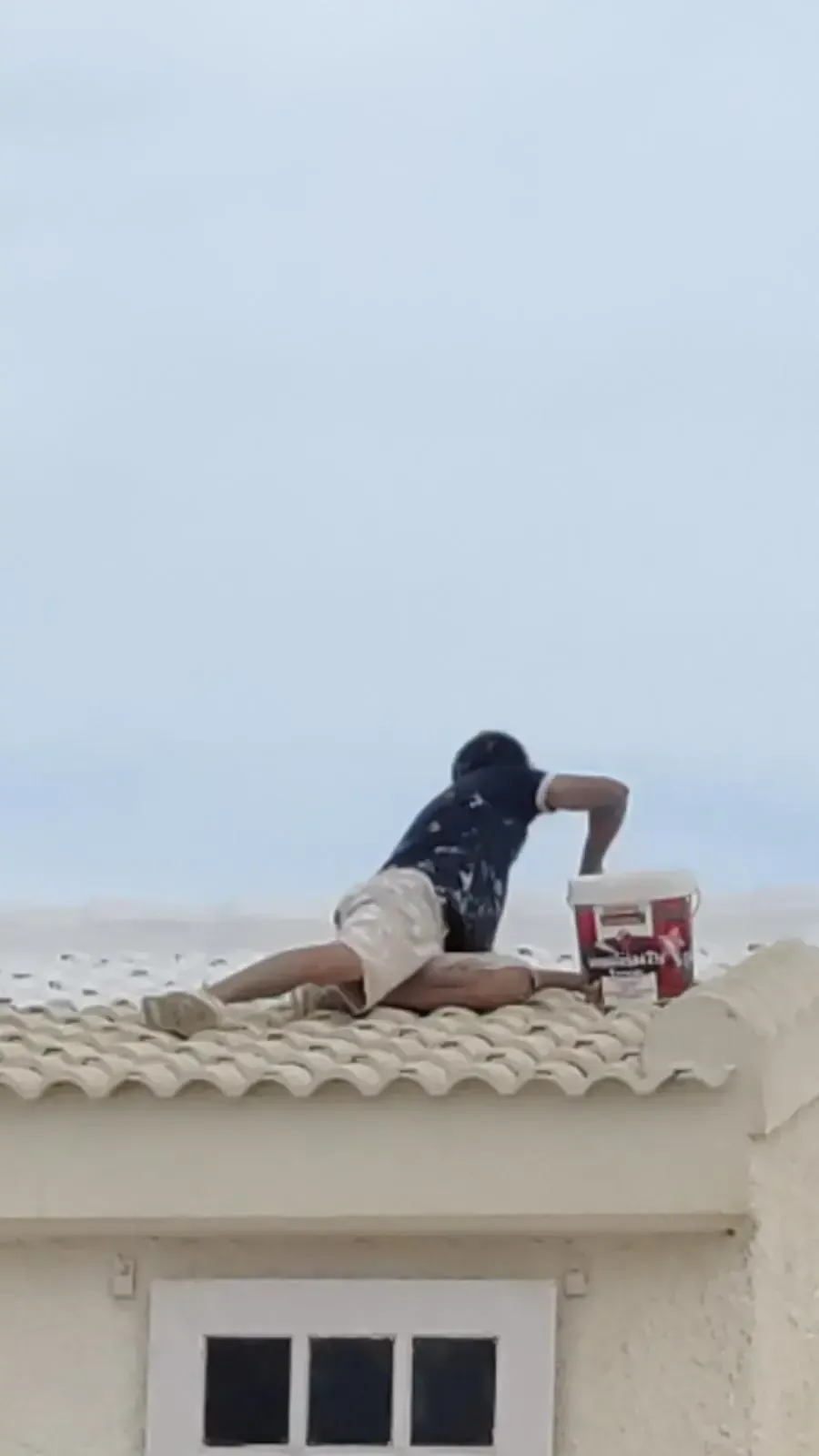 Persona pintando un tejado. Está arrodillada, sosteniendo un cubo de pintura, aplicando pintura a las tejas.