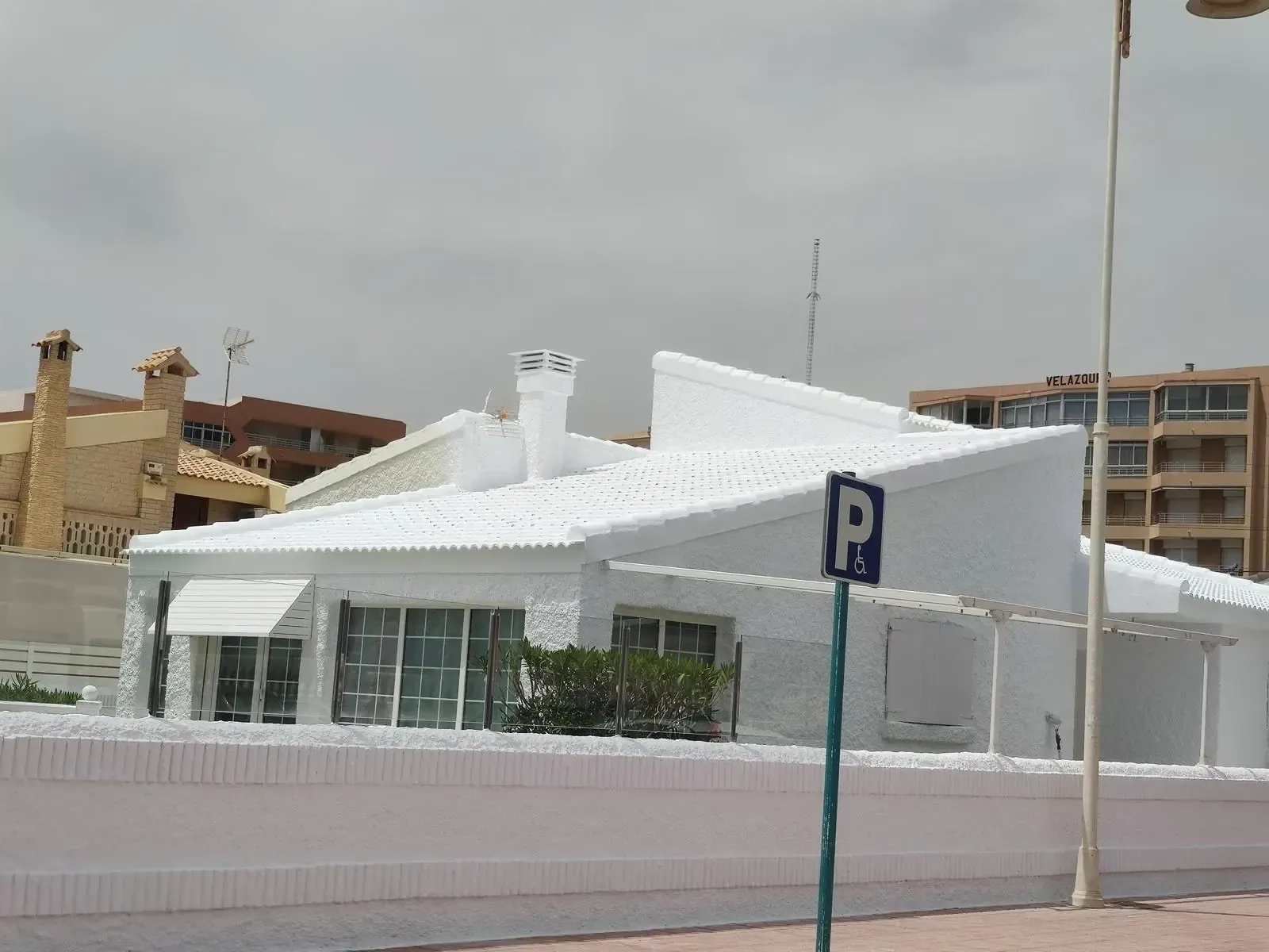 Casa blanca con tejado inclinado, chimenea y un pequeño balcón, junto a un cartel de aparcamiento.
