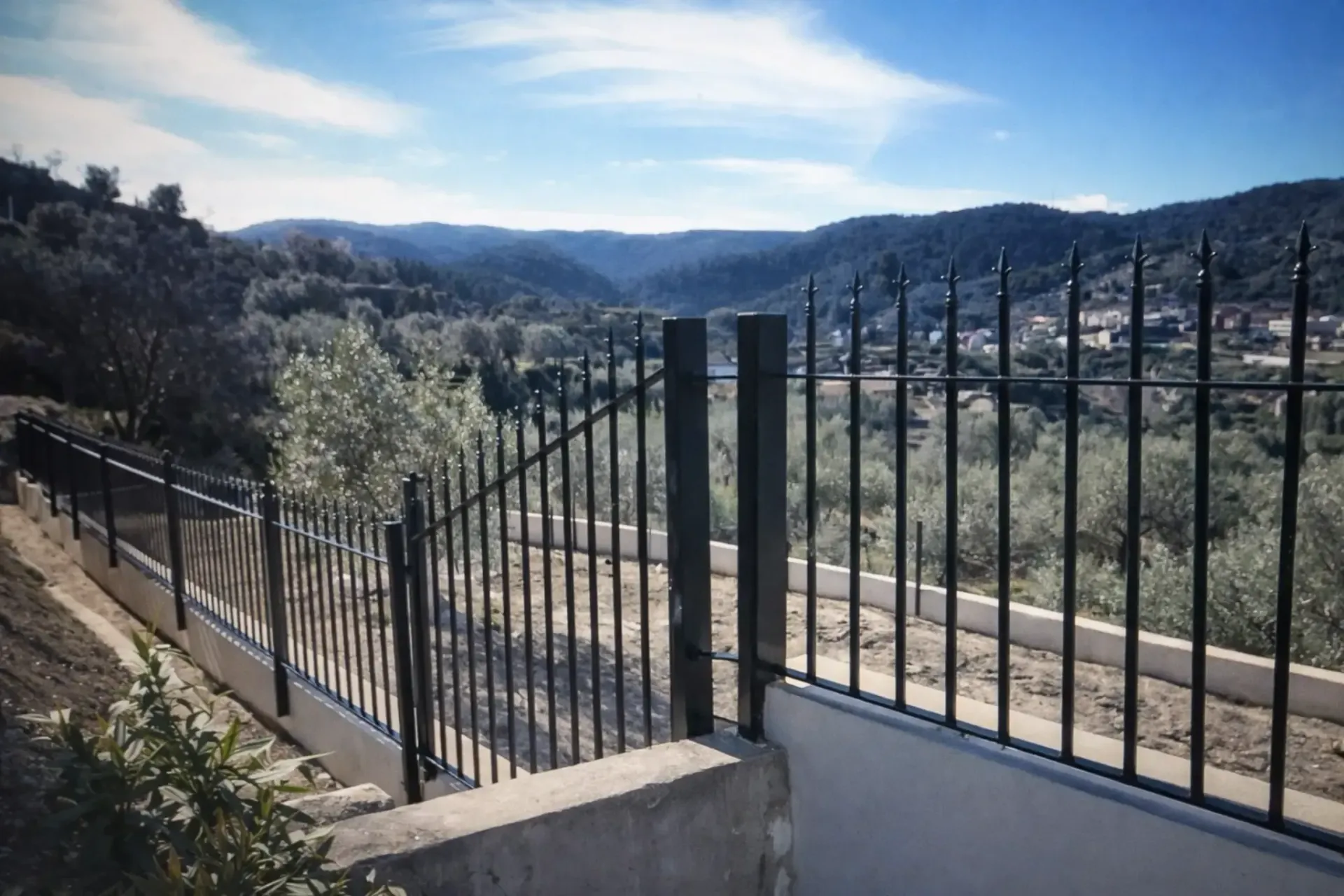 Valla de metal negro con vistas a un valle con colinas onduladas y un cielo azul claro.