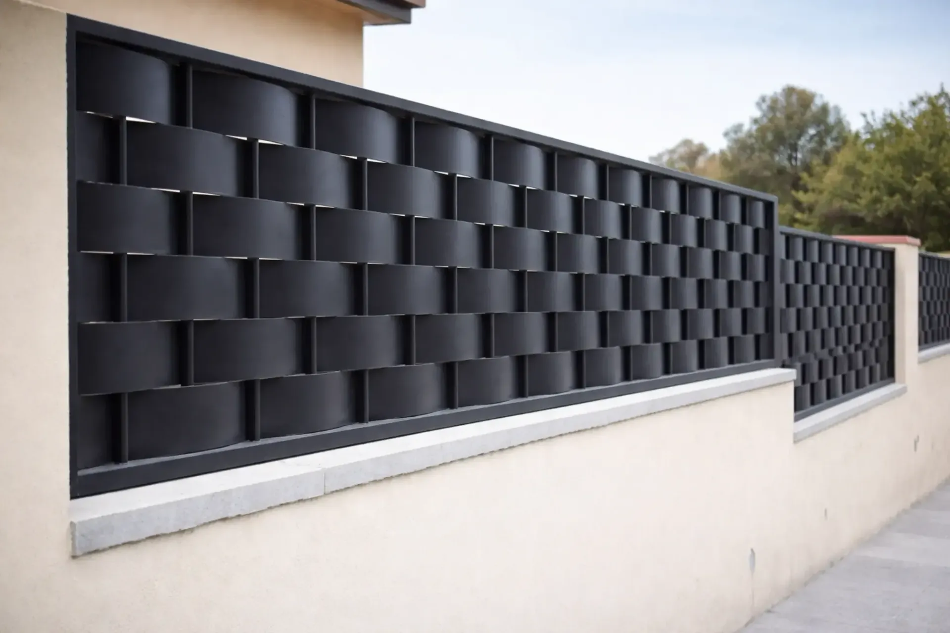 Valla de estilo tejido de color negro sobre una pared de color beige frente a una acera y árboles.