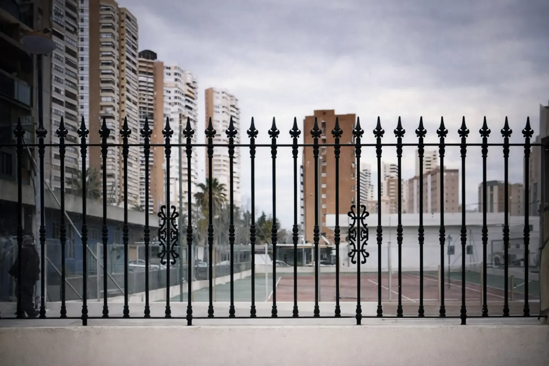 Valla de hierro forjado negro con edificios de apartamentos al fondo, cielo nublado.