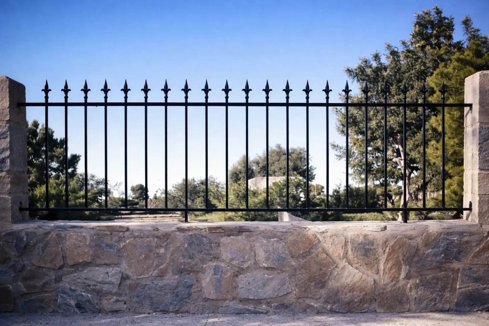 Valla de metal negro sobre un muro de piedra, cielo azul y árboles al fondo.