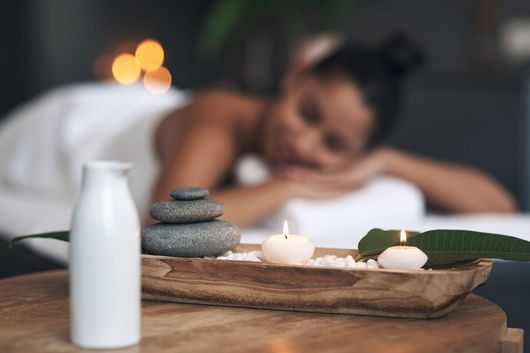Bandeja de spa con velas, piedras apiladas y una botella blanca, con una persona ligeramente desenfocada al fondo.