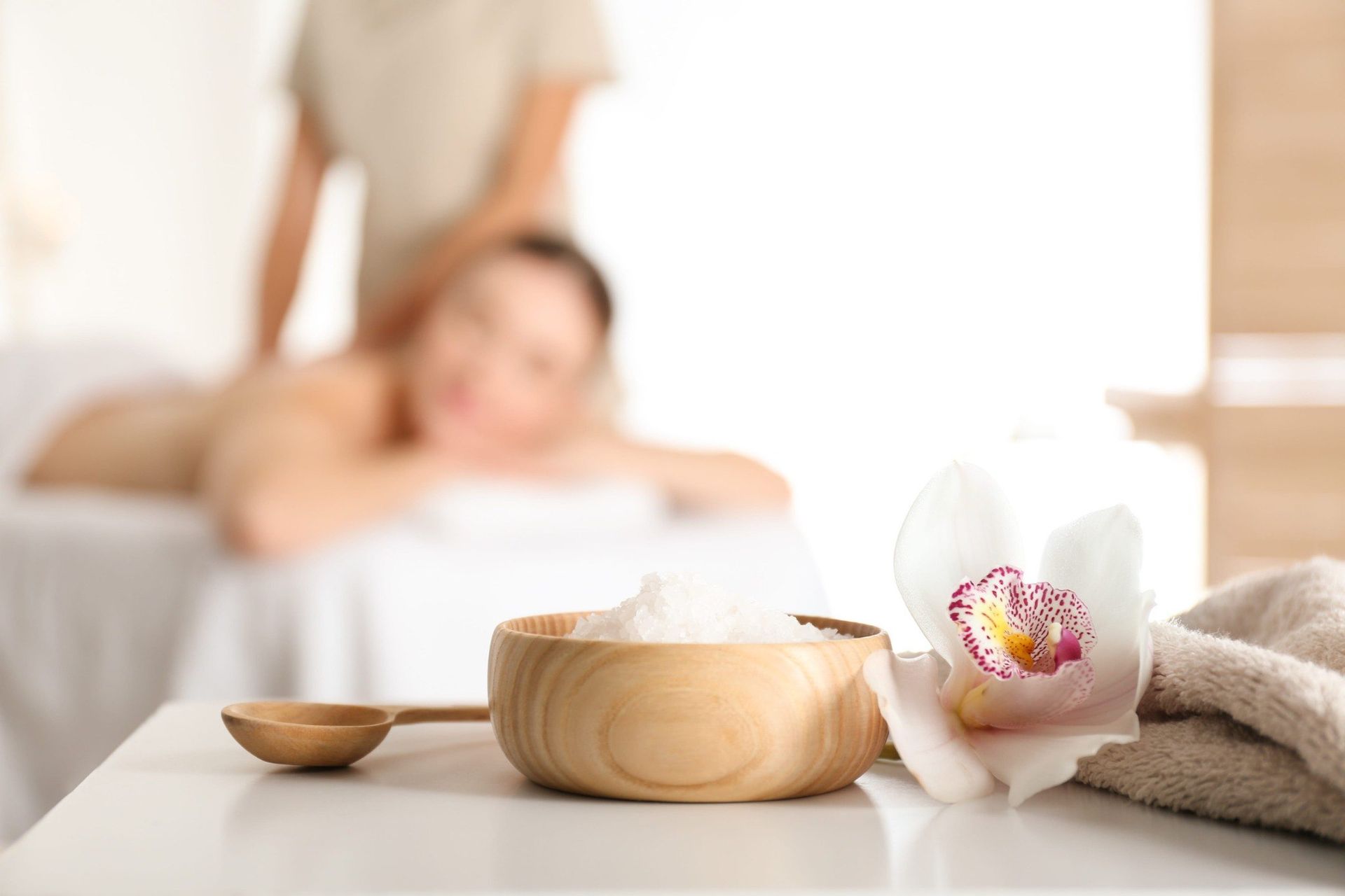 Un cuenco de madera para spa con flores blancas y una toalla sobre una mesa, con una escena de masaje borrosa al fondo.