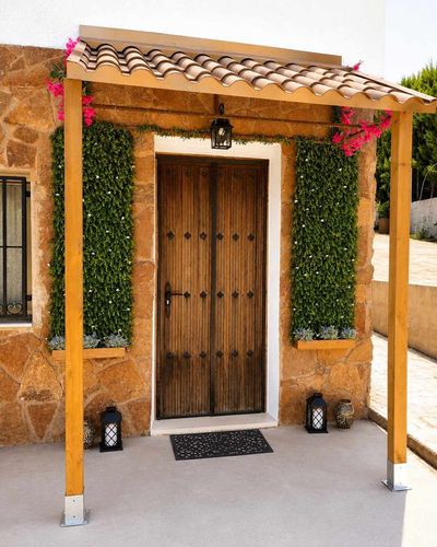 Puerta principal rústica de madera con paredes cubiertas de hiedra, postes amarillos y flores rosas.