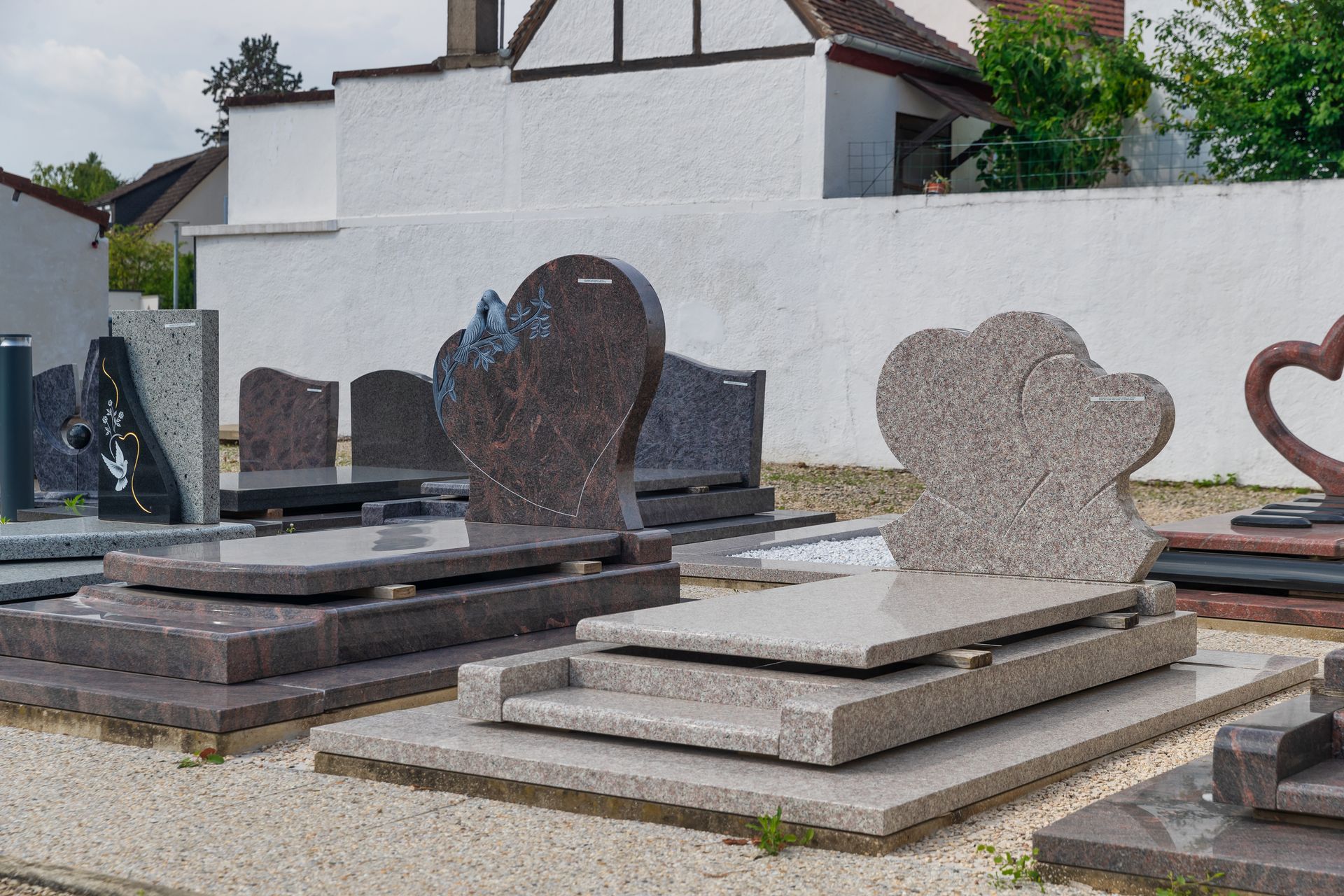 Plusieurs pierres tombales en granit aux formes distinctes se dressent devant un mur blanc, dans un cimetière extérieur.