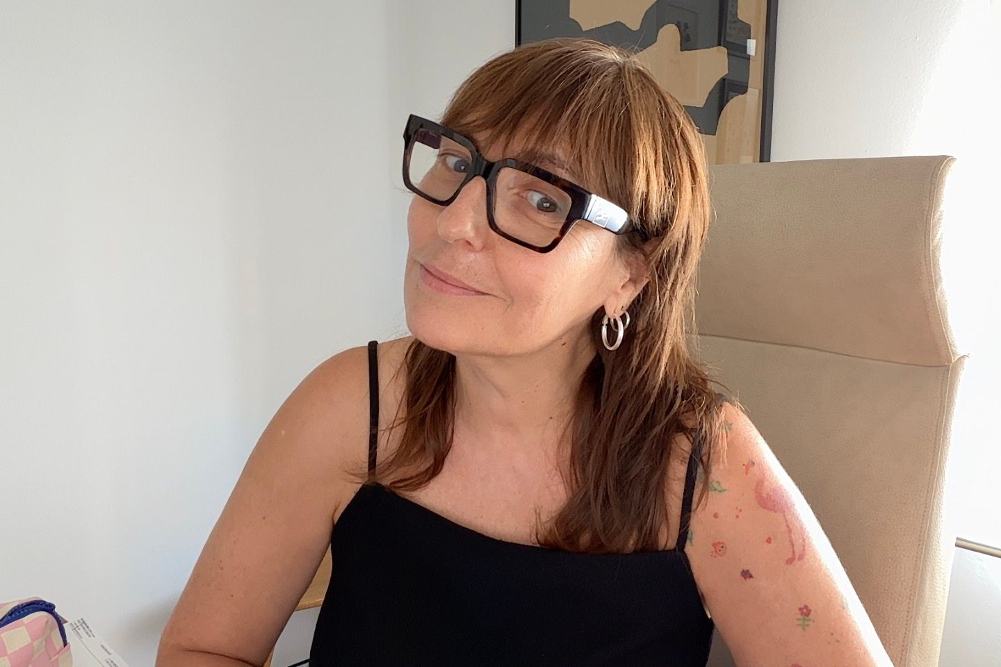 Mujer con gafas sonríe, sentada en una silla. Suéter negro con lunares rojos, pendiente de perla.