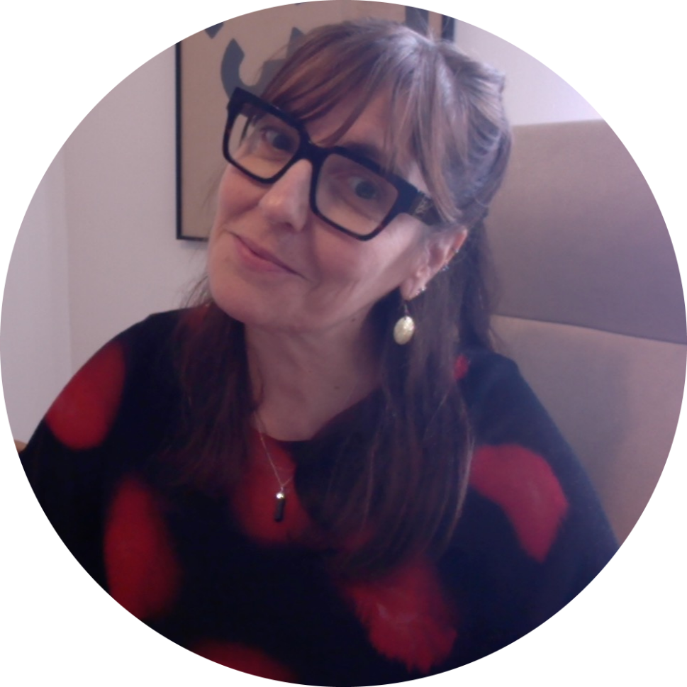 Mujer con gafas sonríe, sentada en una silla. Suéter negro con lunares rojos, pendiente de perla.