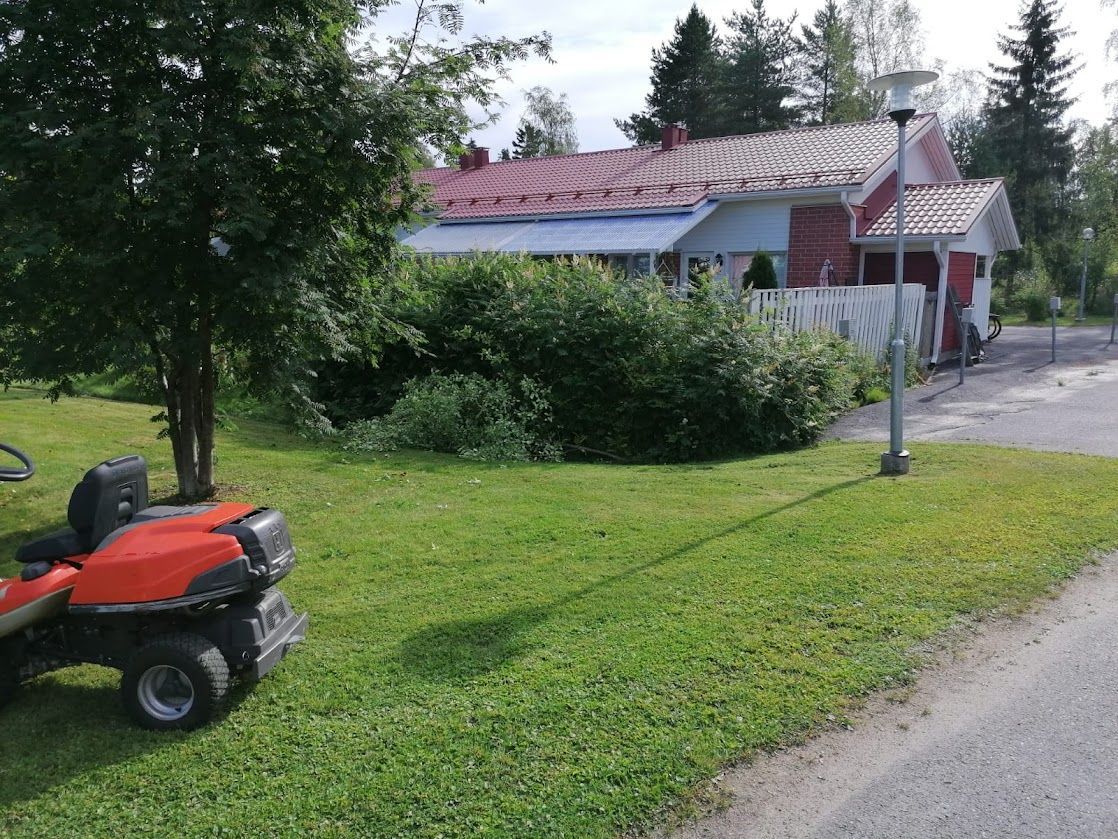 Punainen ruohonleikkuri nurmikolla maaseudun talon ja pensaiden ja puiden täyttämän ajotien vieressä