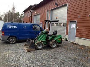 Vihreä traktori etukuormaajalla pysäköitynä sinisen pakettiauton viereen punaisen teollisuusrakennuksen ulkopuolelle.