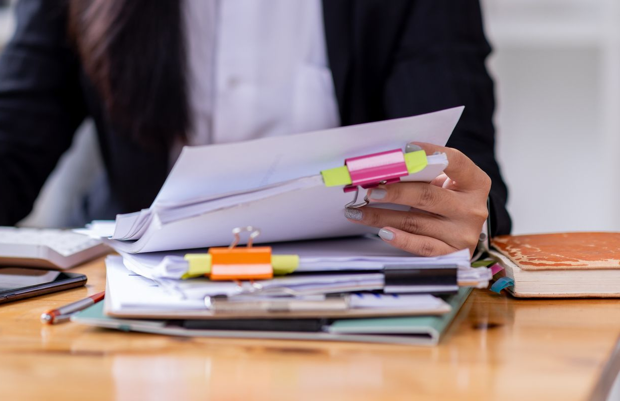 Une personne en blazer, assise à un bureau, trie une pile de papiers maintenus par des trombones.