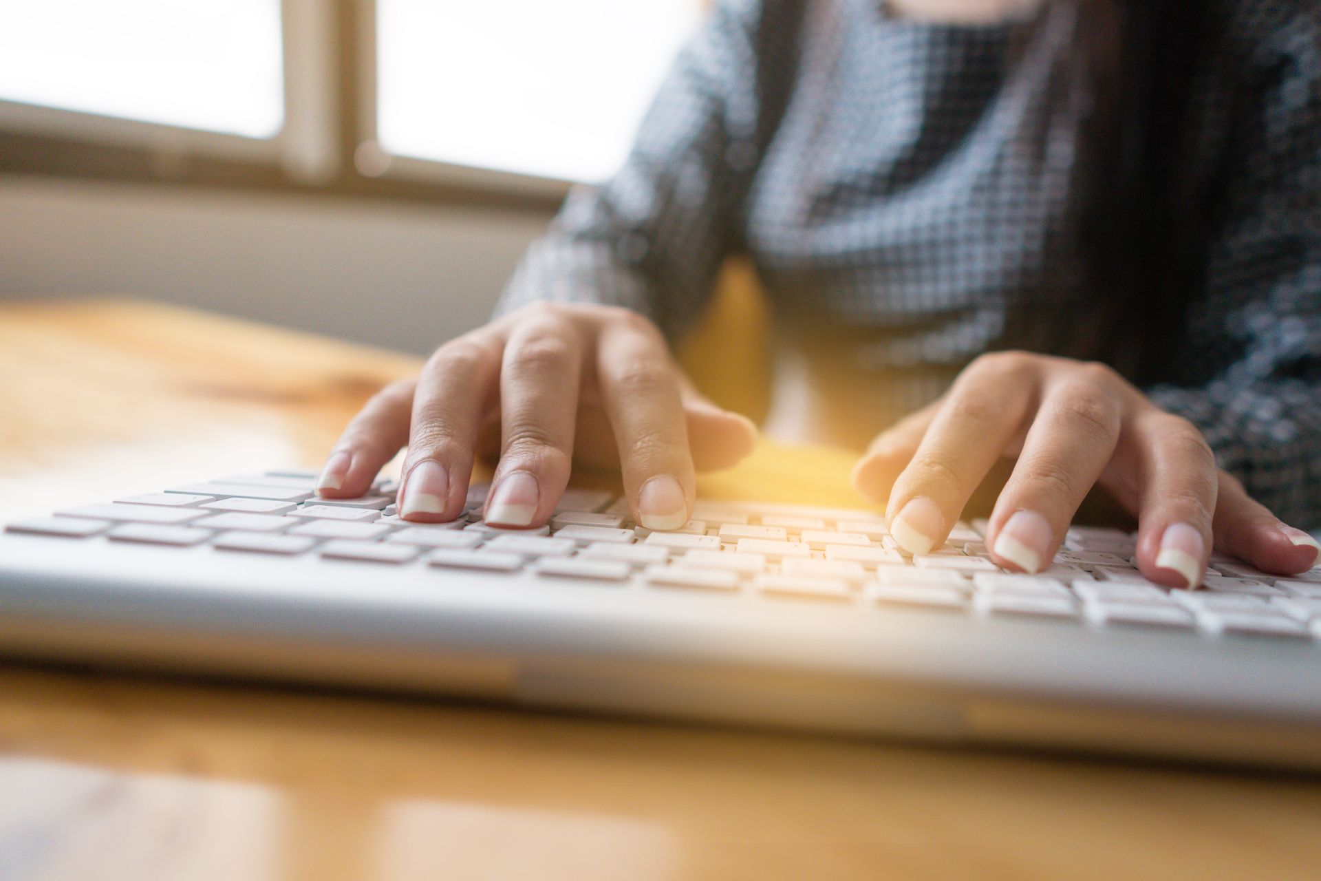 Des mains tapant sur un clavier blanc posé sur un bureau en bois près d'une fenêtre.