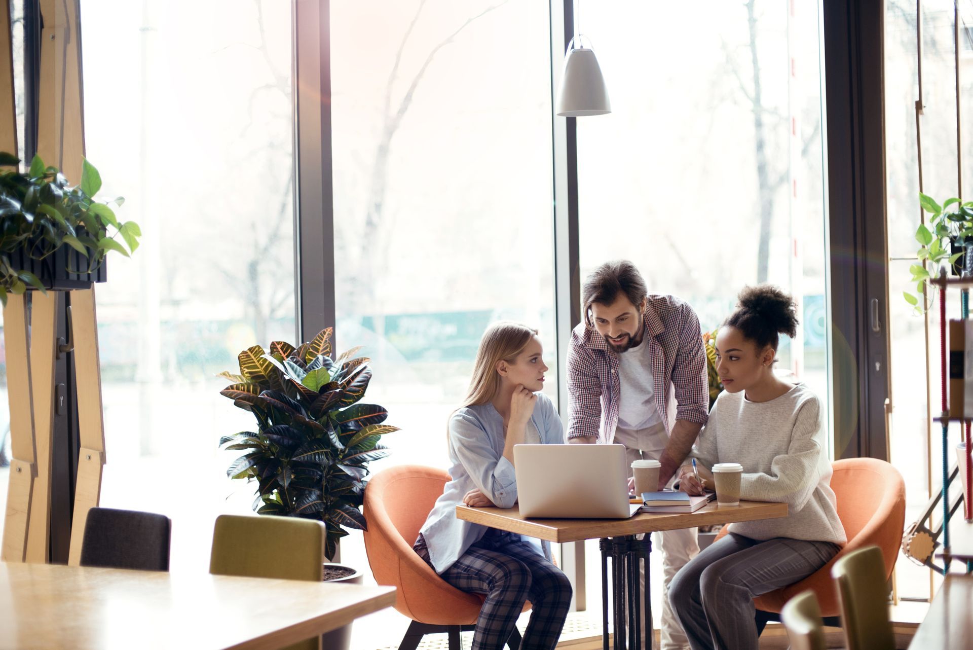 Trois personnes collaborent autour d'un ordinateur portable à une table de café, à la lumière naturelle.